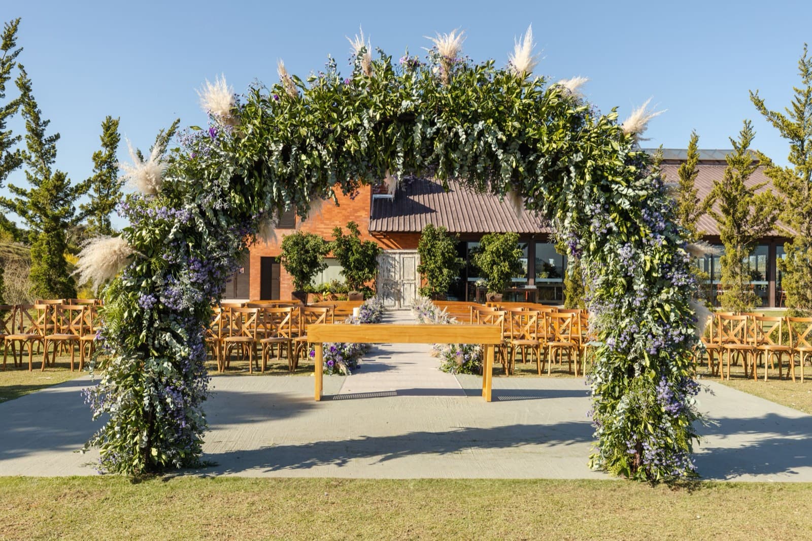 A greenery-and-pampas ceremony arch frames rows of wooden cross-back chairs in a sunny courtyard