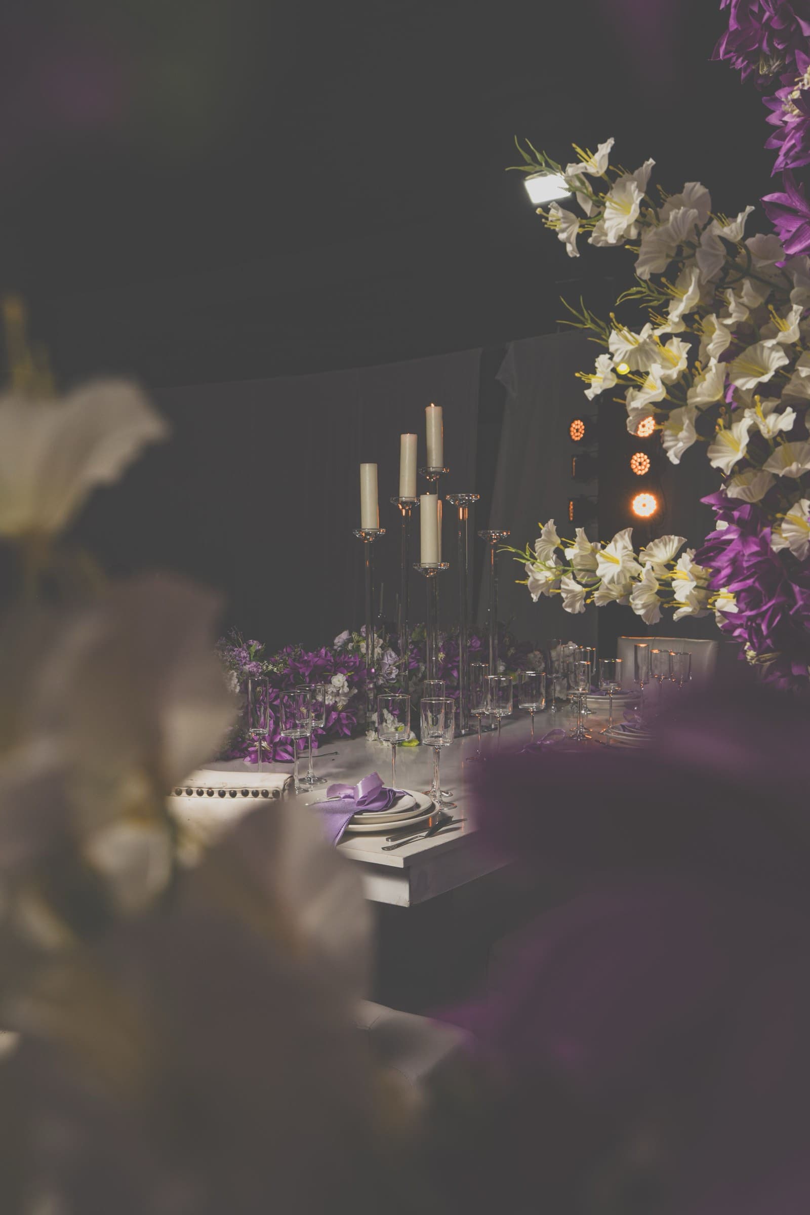 Tall taper candles rising above purple and white florals along a dimly lit head table