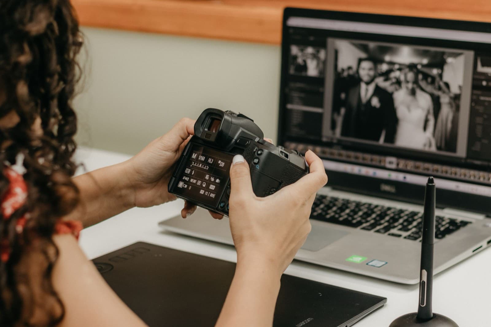 A photographer checks settings on a camera’s rear screen beside a laptop showing a black-and-white wedding portrait