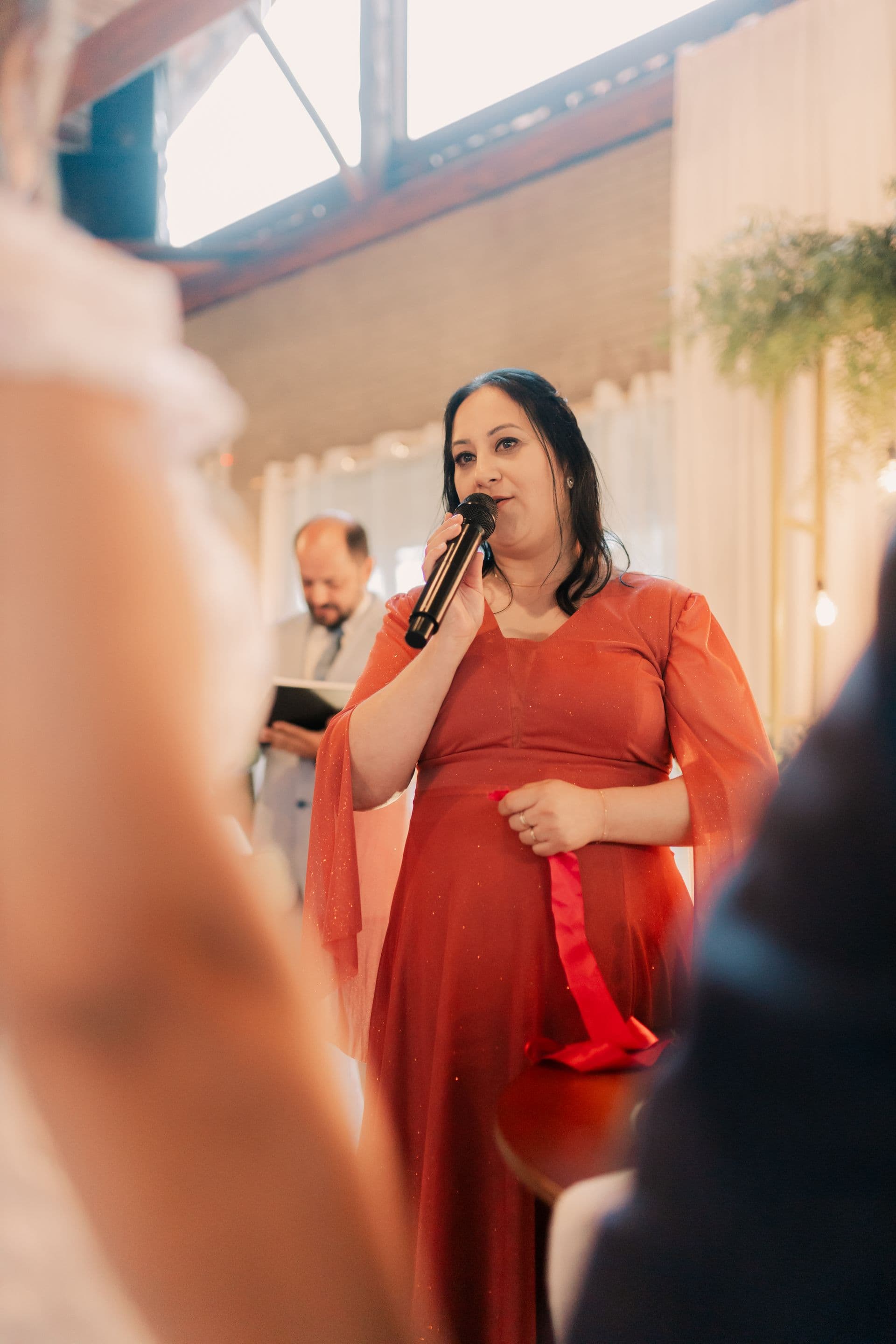 A woman in a flowing red dress speaks into a microphone at a warmly lit indoor ceremony, with the officiant standing behind her