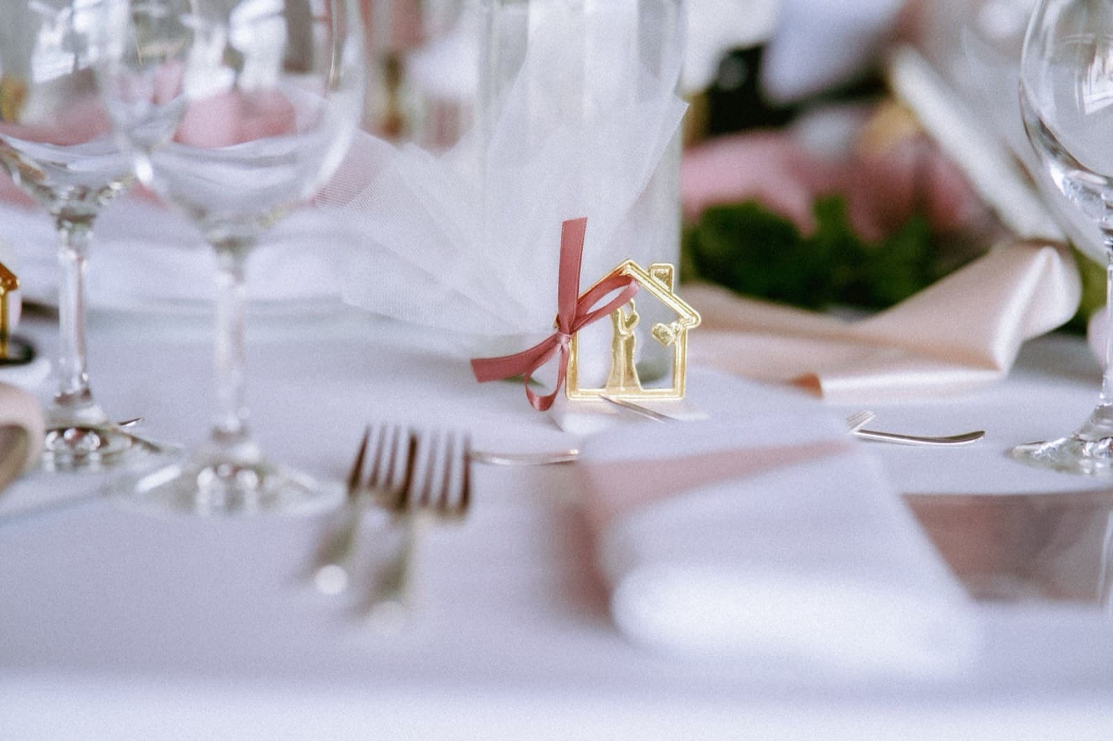 A gold house-shaped wedding favor tied with a rose ribbon among forks, folded napkins, and glasses on a white tablecloth