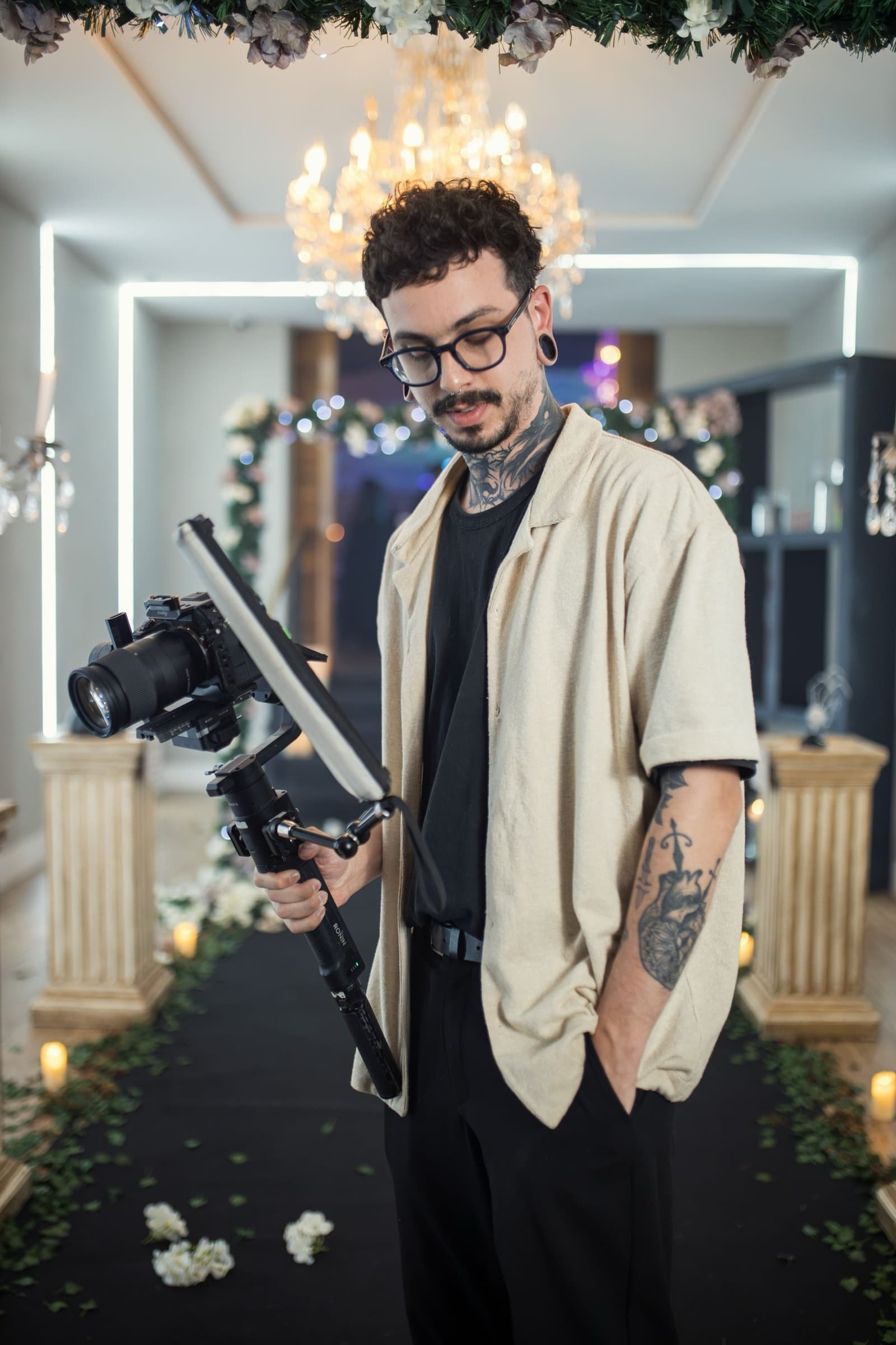 A videographer steadies a gimbal-mounted camera and light beneath a chandelier and greenery garland at a wedding venue