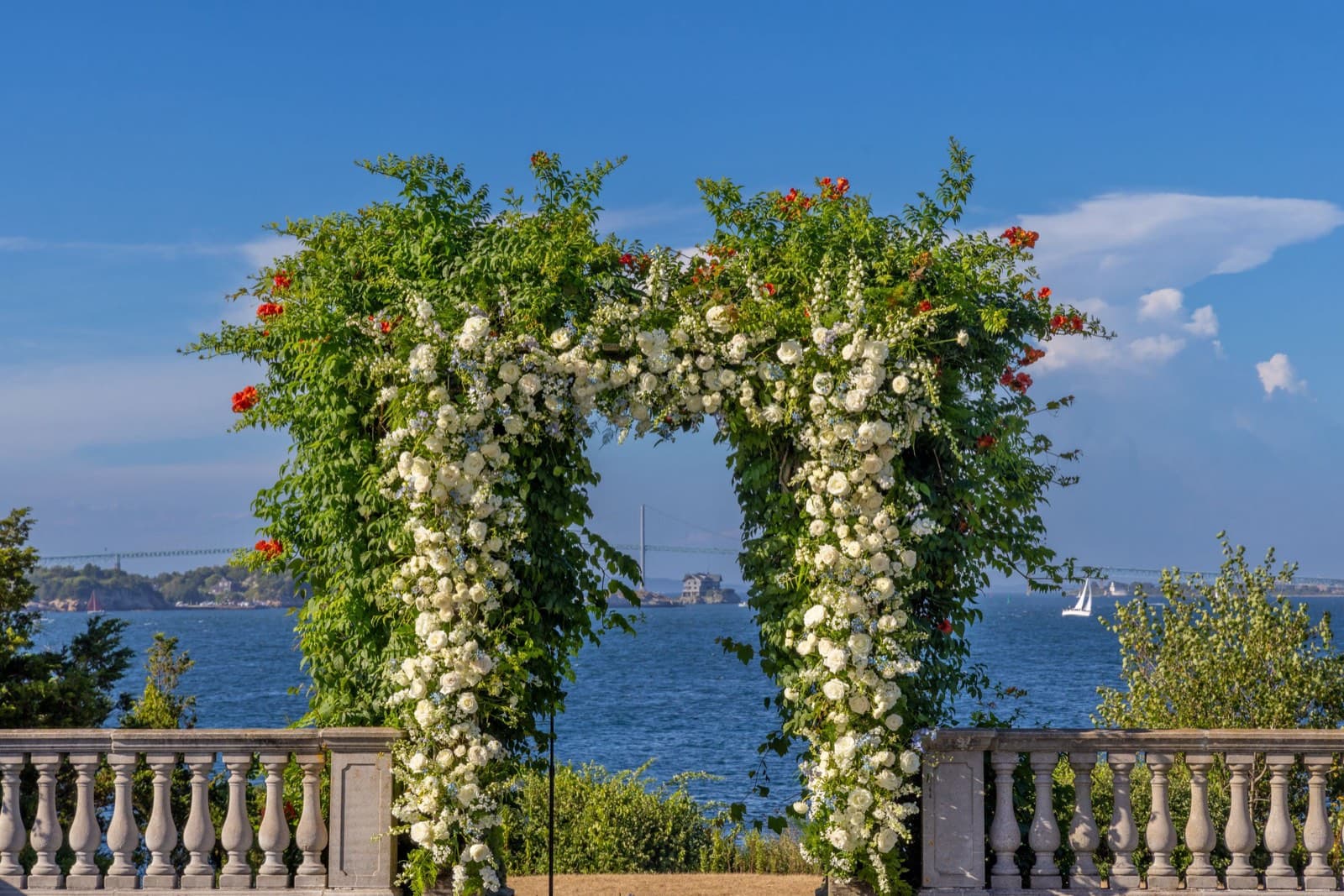 A rose-covered ceremony arch on a stone balustrade terrace overlooking a bay with a distant bridge and sailboat