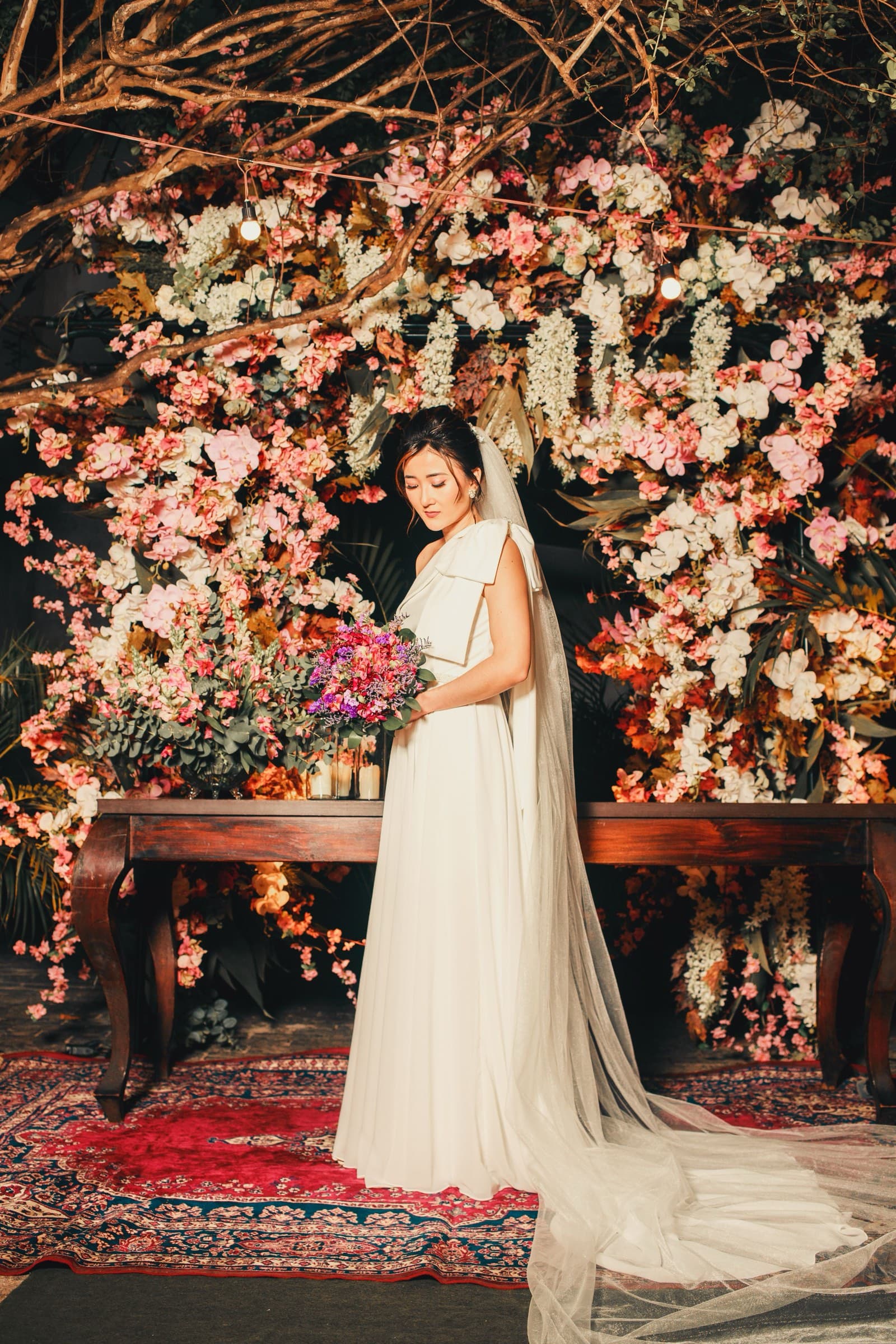 A bride with a long veil holds her bouquet before a wall of pink and white blooms
