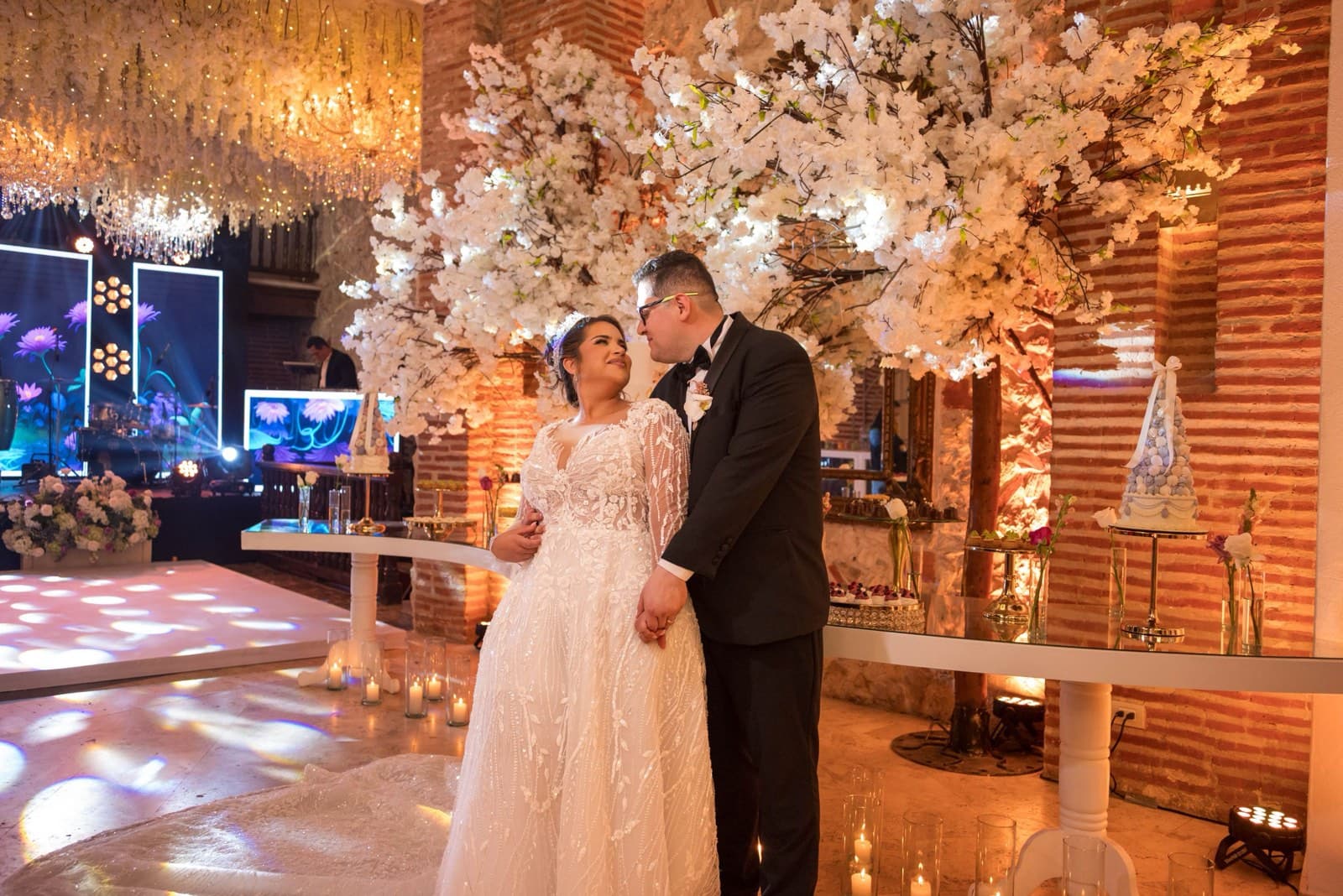A bride and groom hold hands beside white blossom trees and a candlelit dessert display at their reception