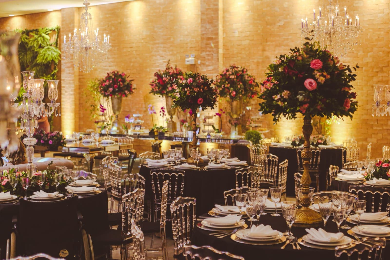 Crystal chandeliers and towering rose centerpieces on gold stands above dark-linen tables in a brick-walled ballroom