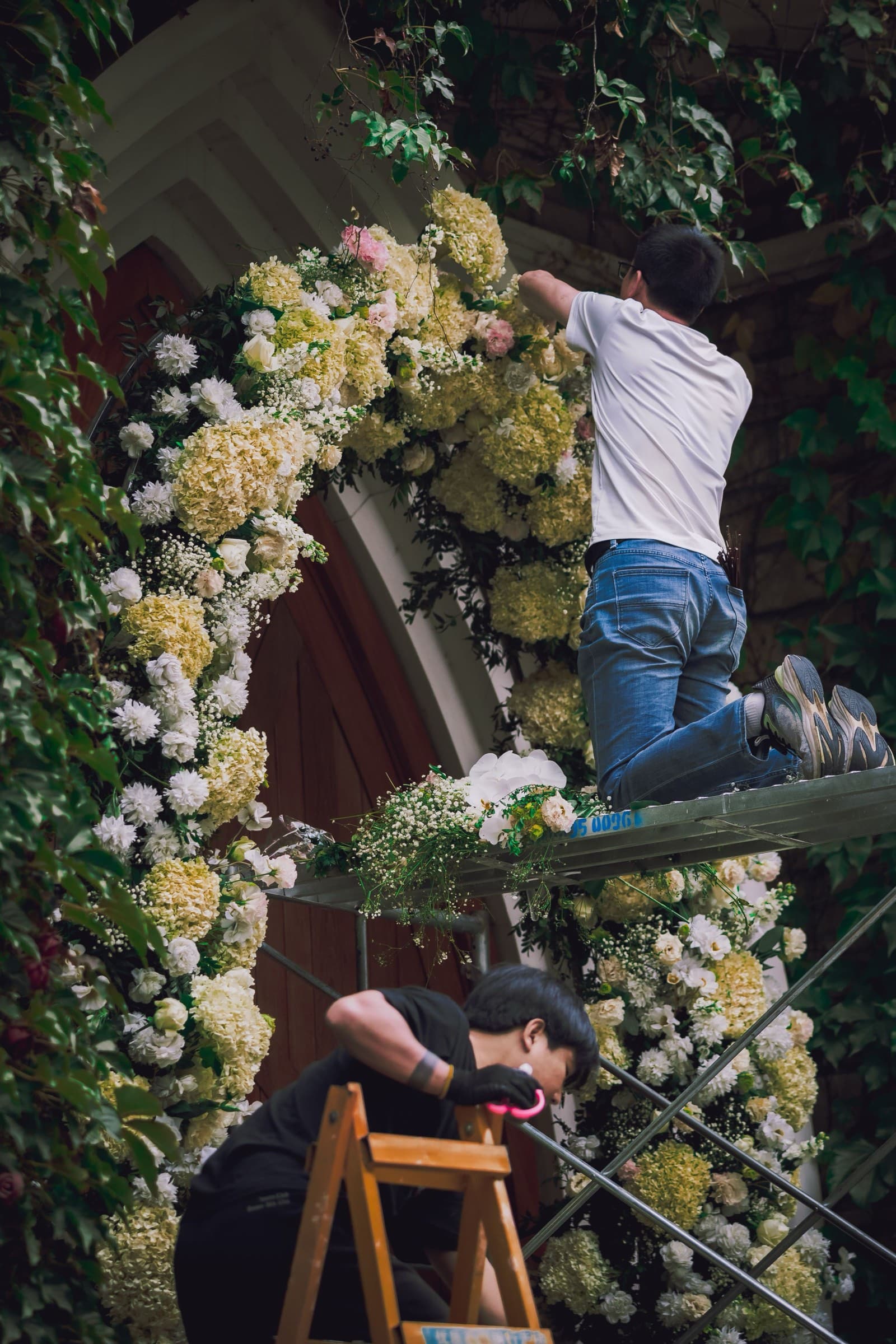 Two florists on ladders dress a ceremony arch with white hydrangeas above a chapel doorway