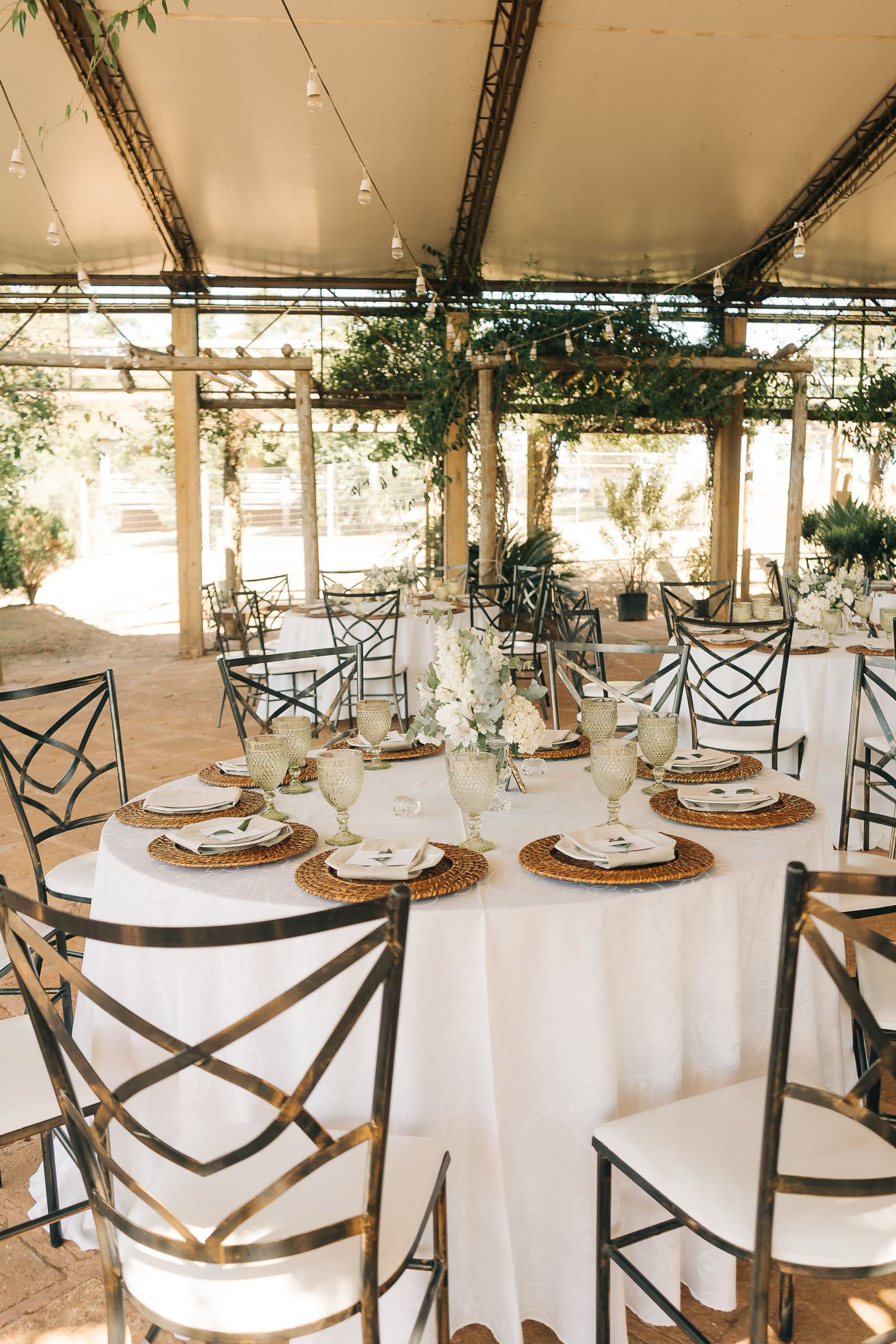 A round table with woven chargers, textured green goblets, and white florals under a vine-covered pergola strung with lights