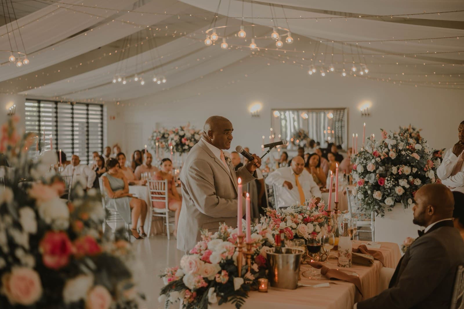 A speaker addresses seated wedding guests beneath draped fabric and string lights, flanked by rose arrangements and pink taper candles