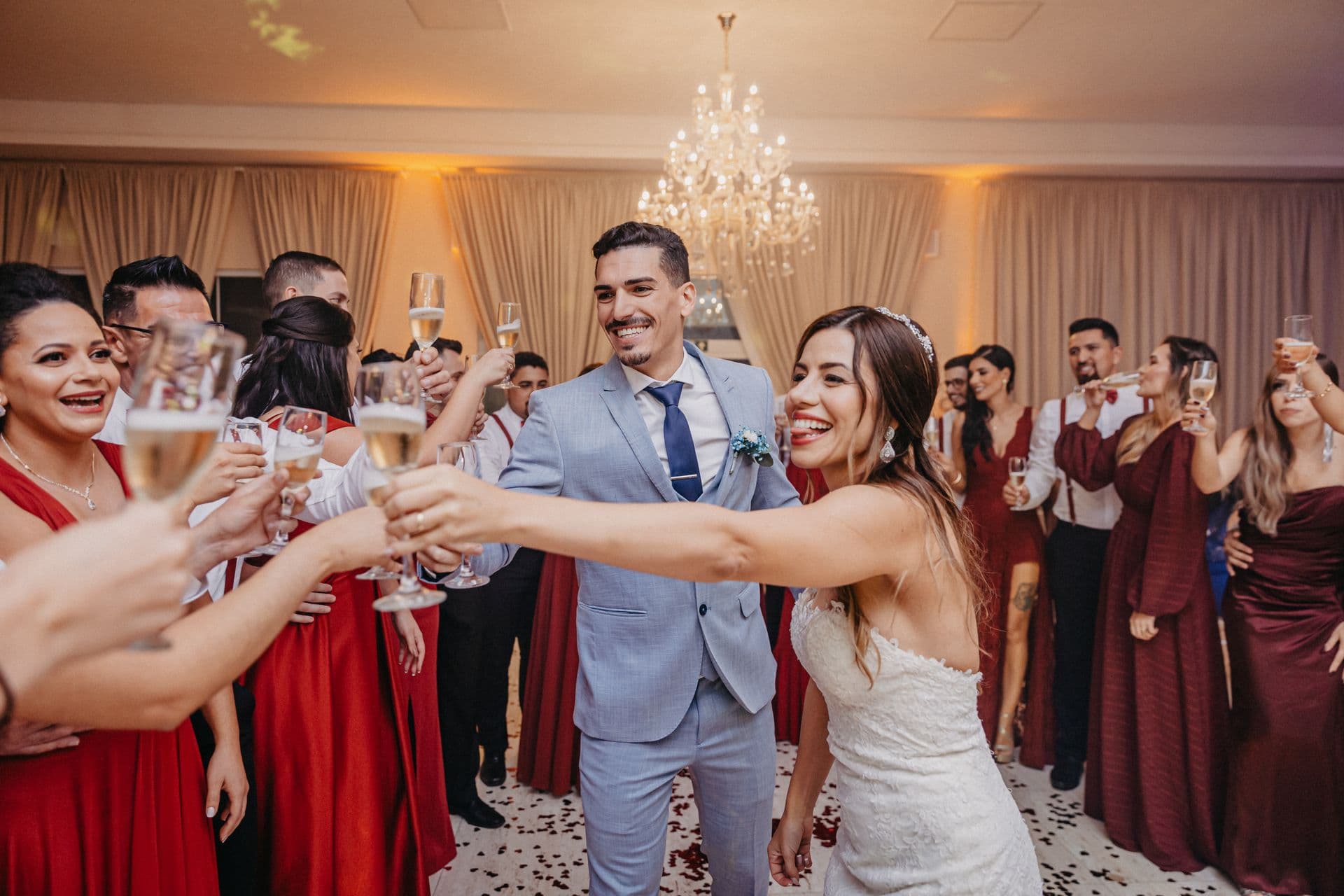 Newlyweds and guests in burgundy raise champagne flutes in a toast beneath a chandelier at a warmly lit reception