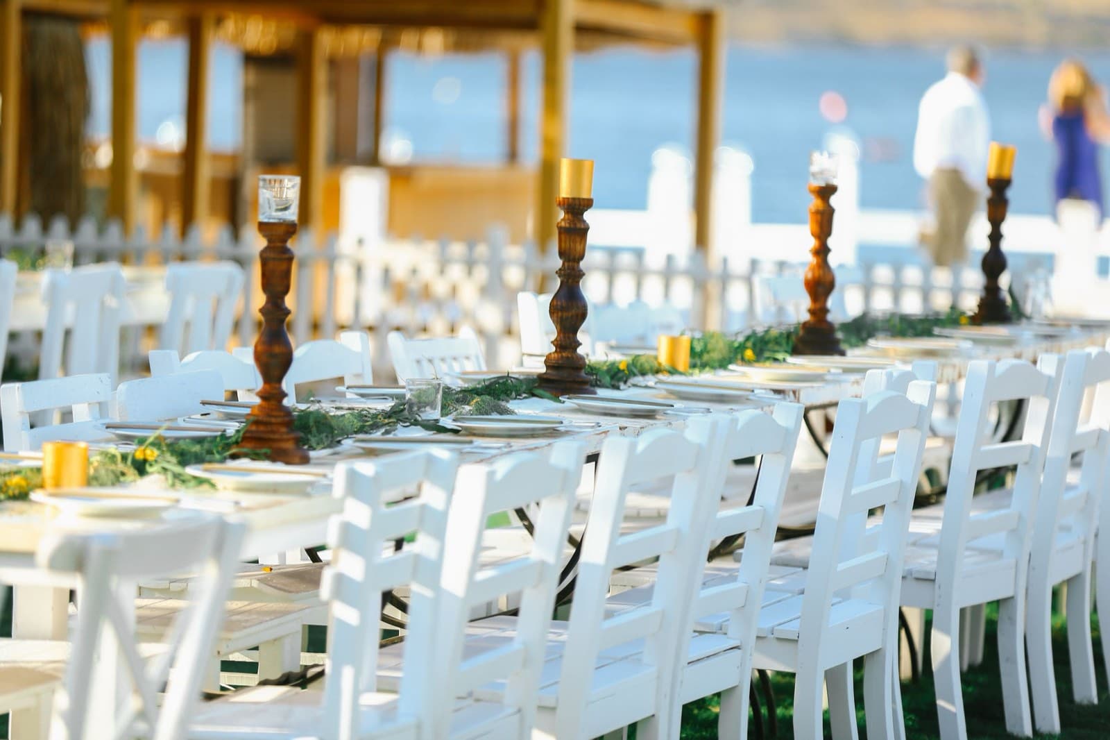 Long white banquet tables with wooden candlesticks and greenery runners set on a lawn overlooking the water