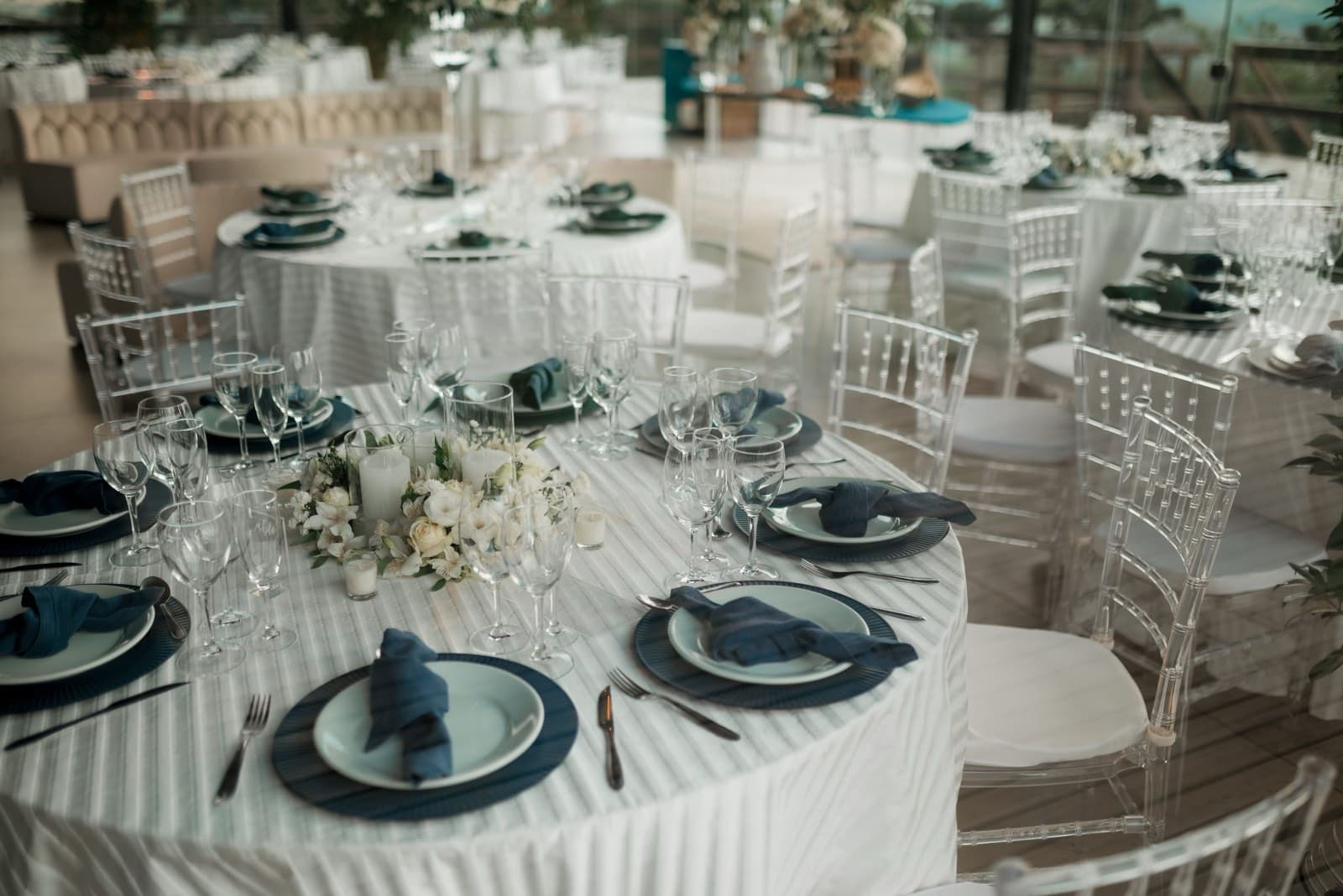 Round tables set with navy napkins, patterned chargers, and white flower centerpieces beside clear chiavari chairs in a glass-walled hall