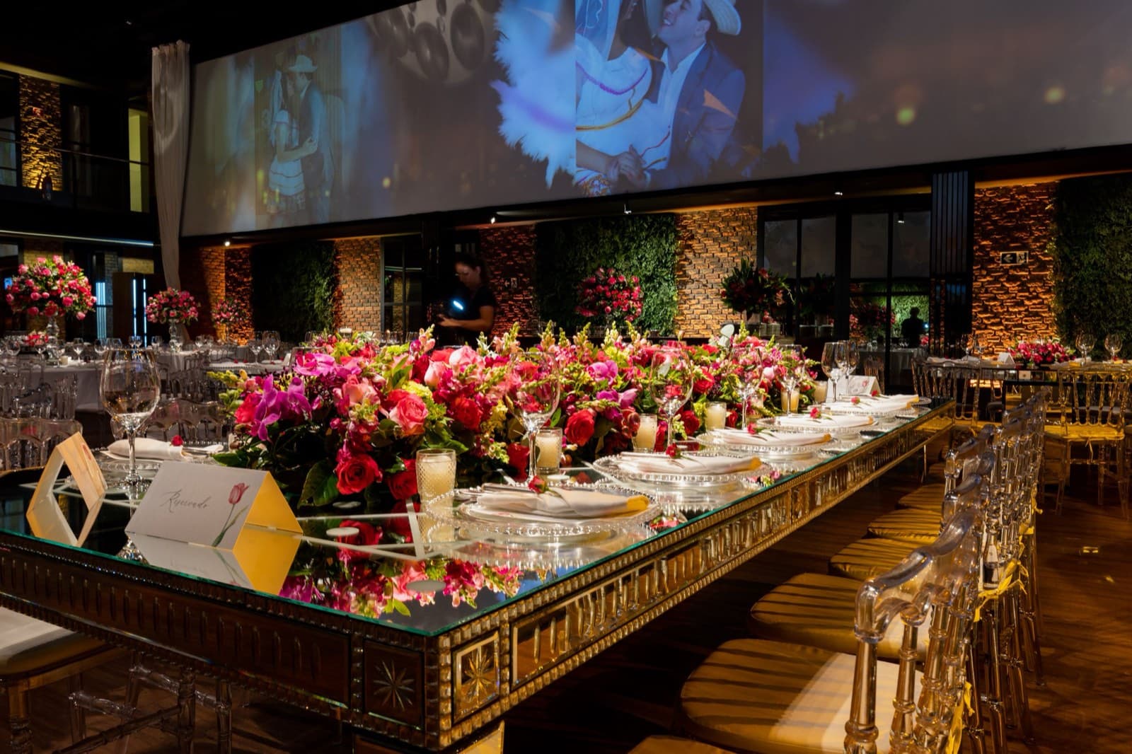 A mirrored banquet table set with lush pink and red florals, glassware, and place cards in a warmly lit venue